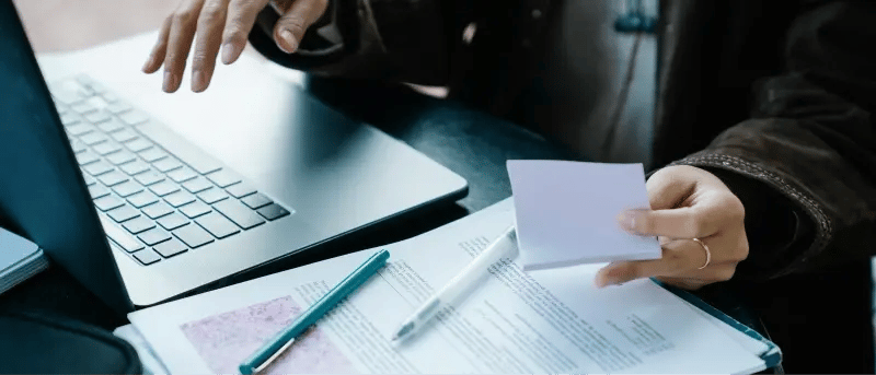 Person using a laptop at a table with papers and a pen, representing technical documentation.